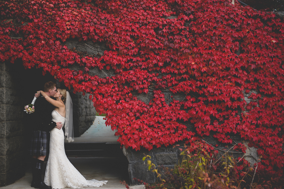 Kiss under vivid fall red leaves outside rossland courtyard, by Nelson BC Kootenay wedding photographer Emilee Zaitsoff of Electrify Photography